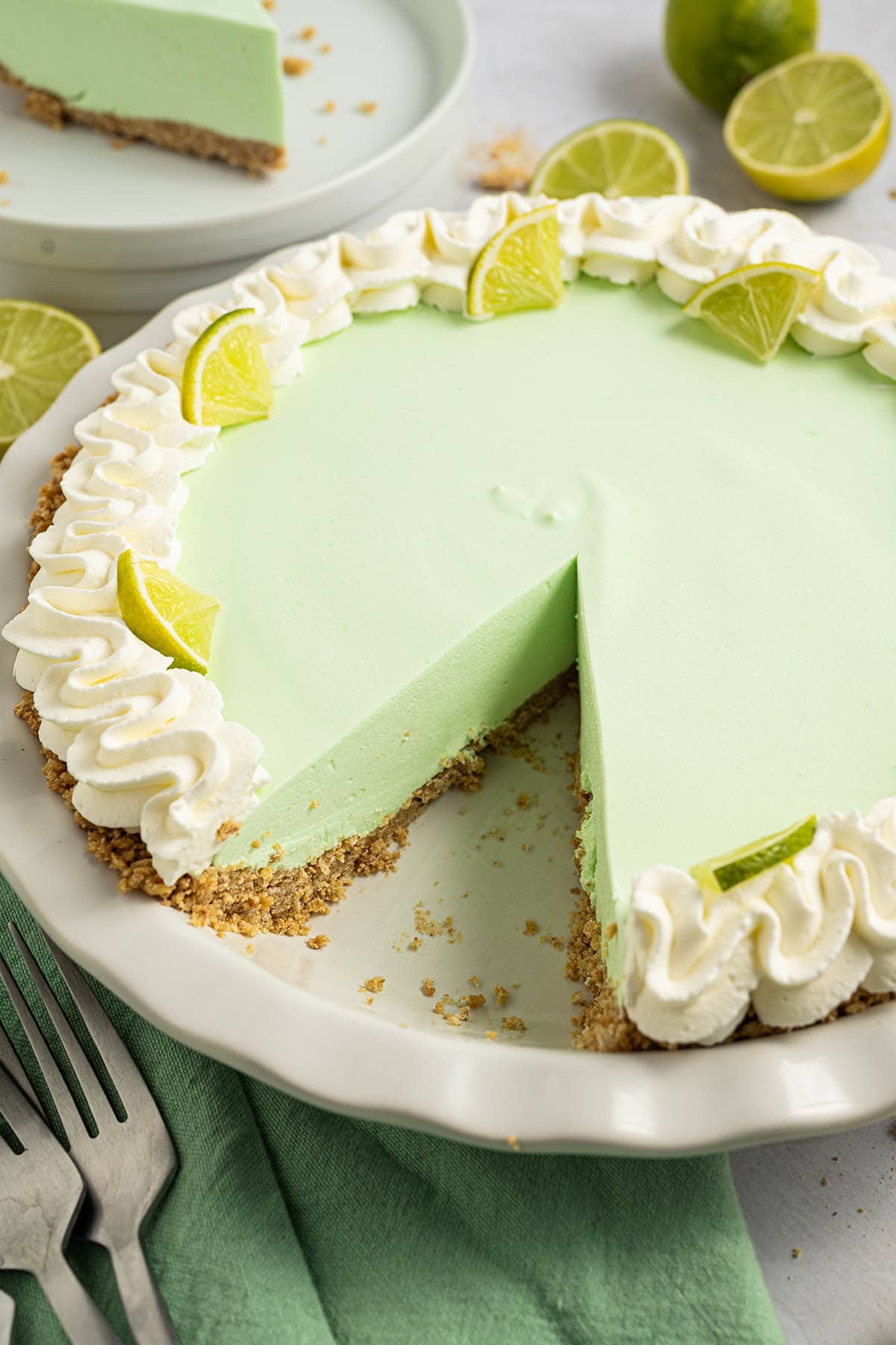 A full lime Jello pie with a graham cracker crust and a slice missing, decorated with whipped cream and lime wedges around the edge.