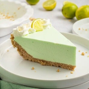A slice of creamy lime Jello pie with a graham cracker crust, topped with whipped cream and a lime wedge, served on a white plate.