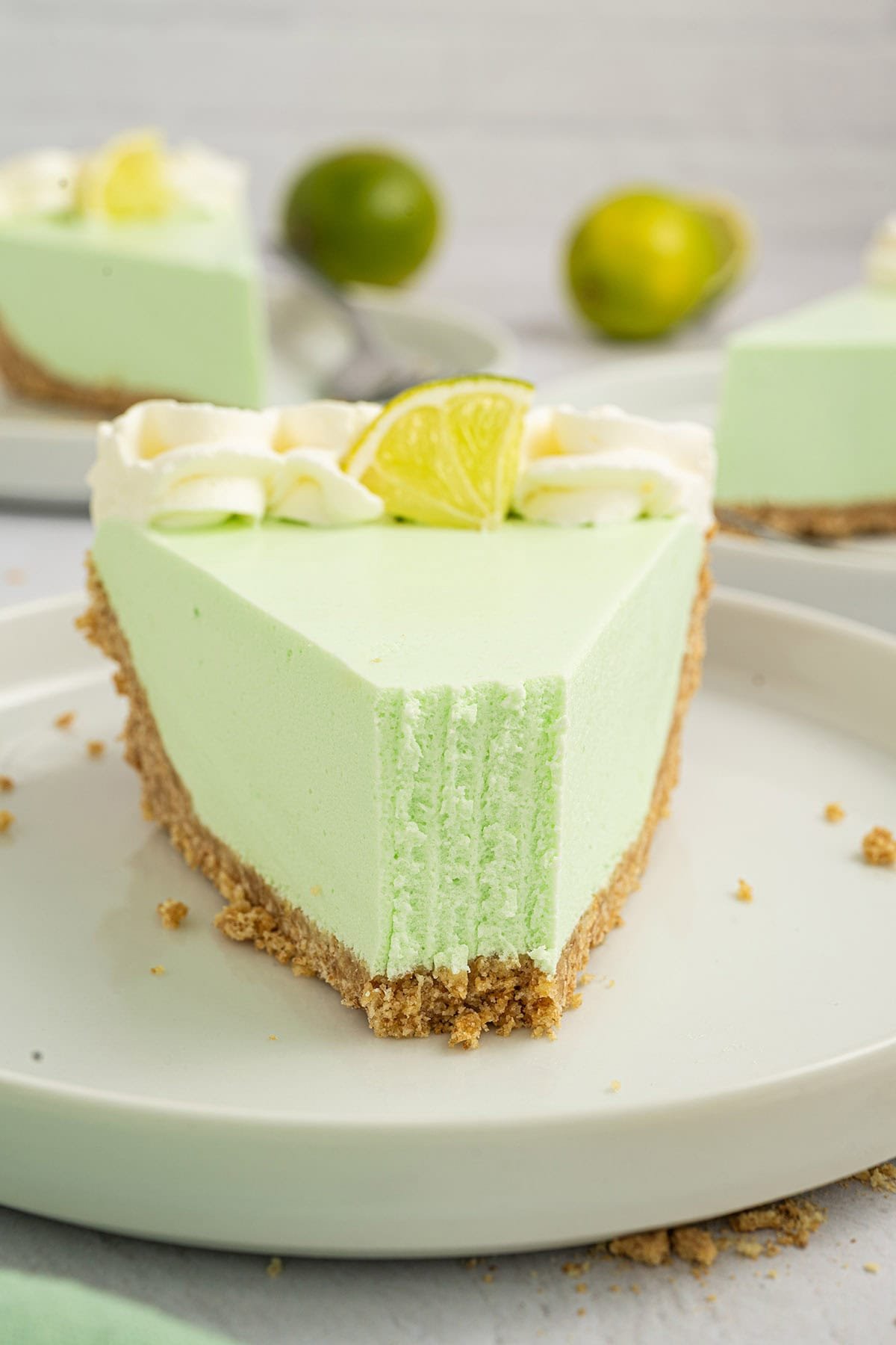 A slice of creamy lime Jello pie with a graham cracker crust and a bite taken out, topped with whipped cream and a lime wedge, served on a white plate.