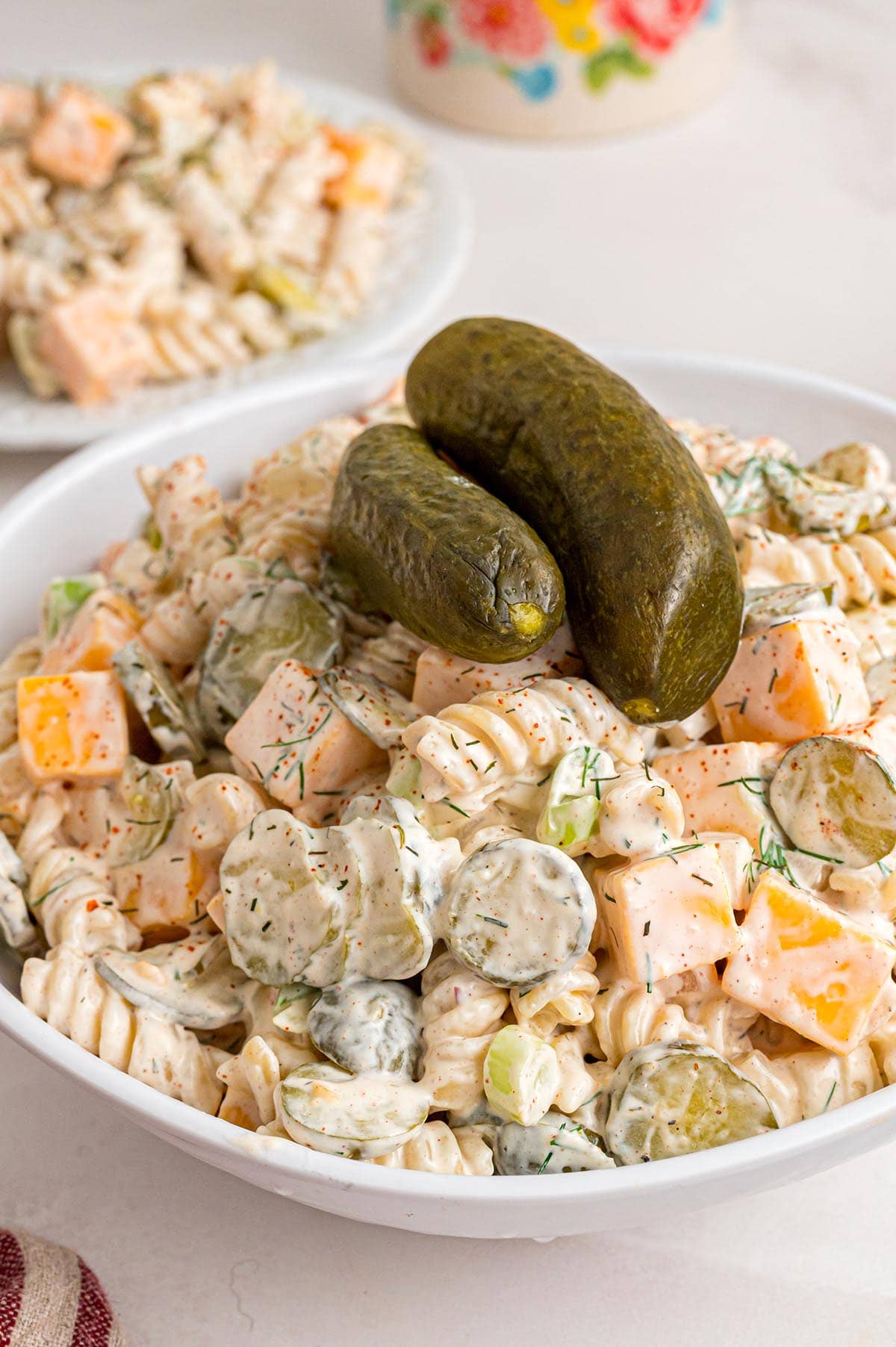 Creamy dill pickle pasta salad in a white bowl with rotini, cheddar cubes, sliced pickles, and whole pickles on top.