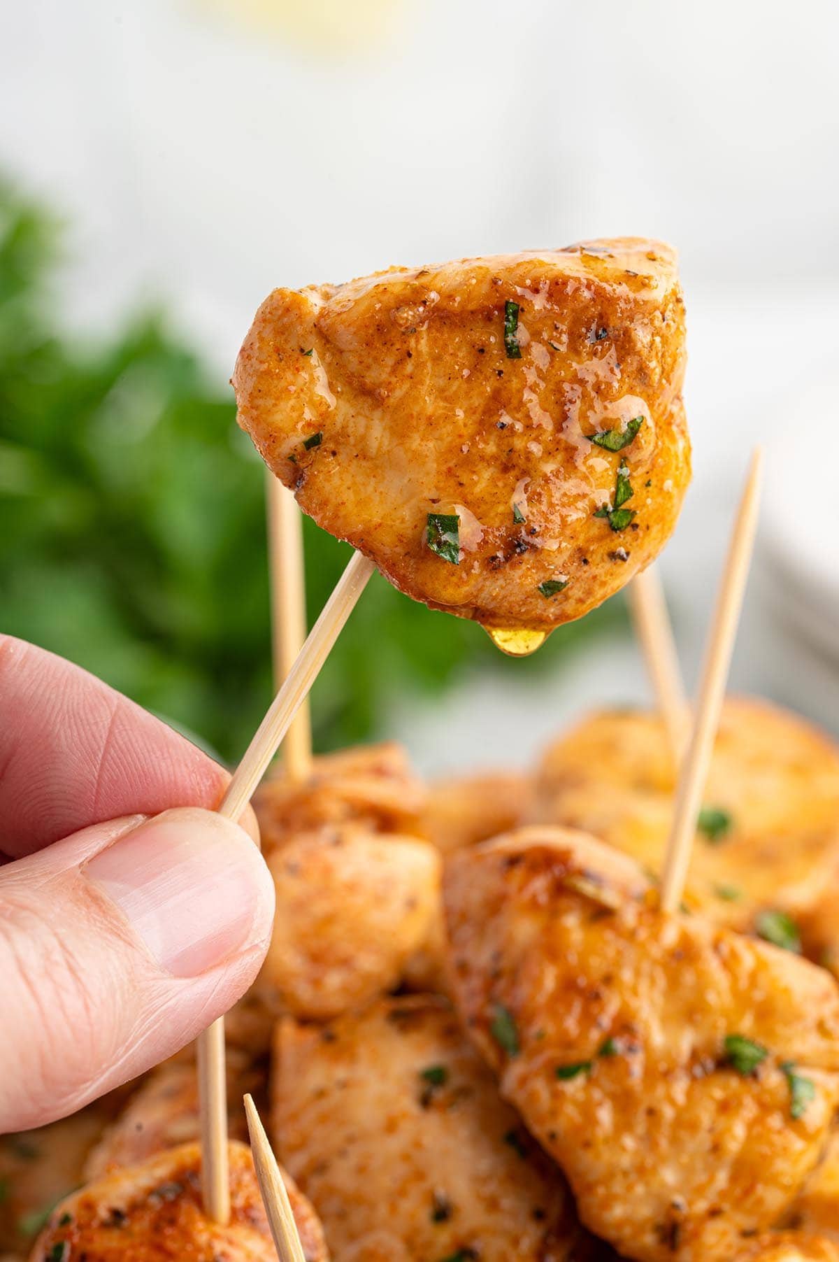 Close-up of a hand holding a juicy garlic butter chicken bite on a toothpick, dripping with sauce and sprinkled with herbs.