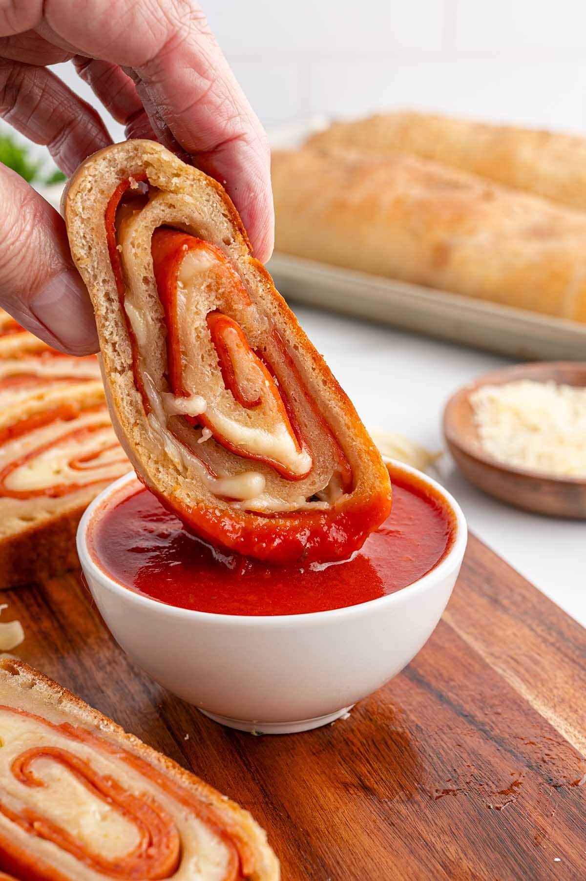 A hand holding a pepperoni roll dipped into a bowl of red marinara sauce.