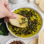 A hand dipping a piece of bread into olive oil with herbs and spices.