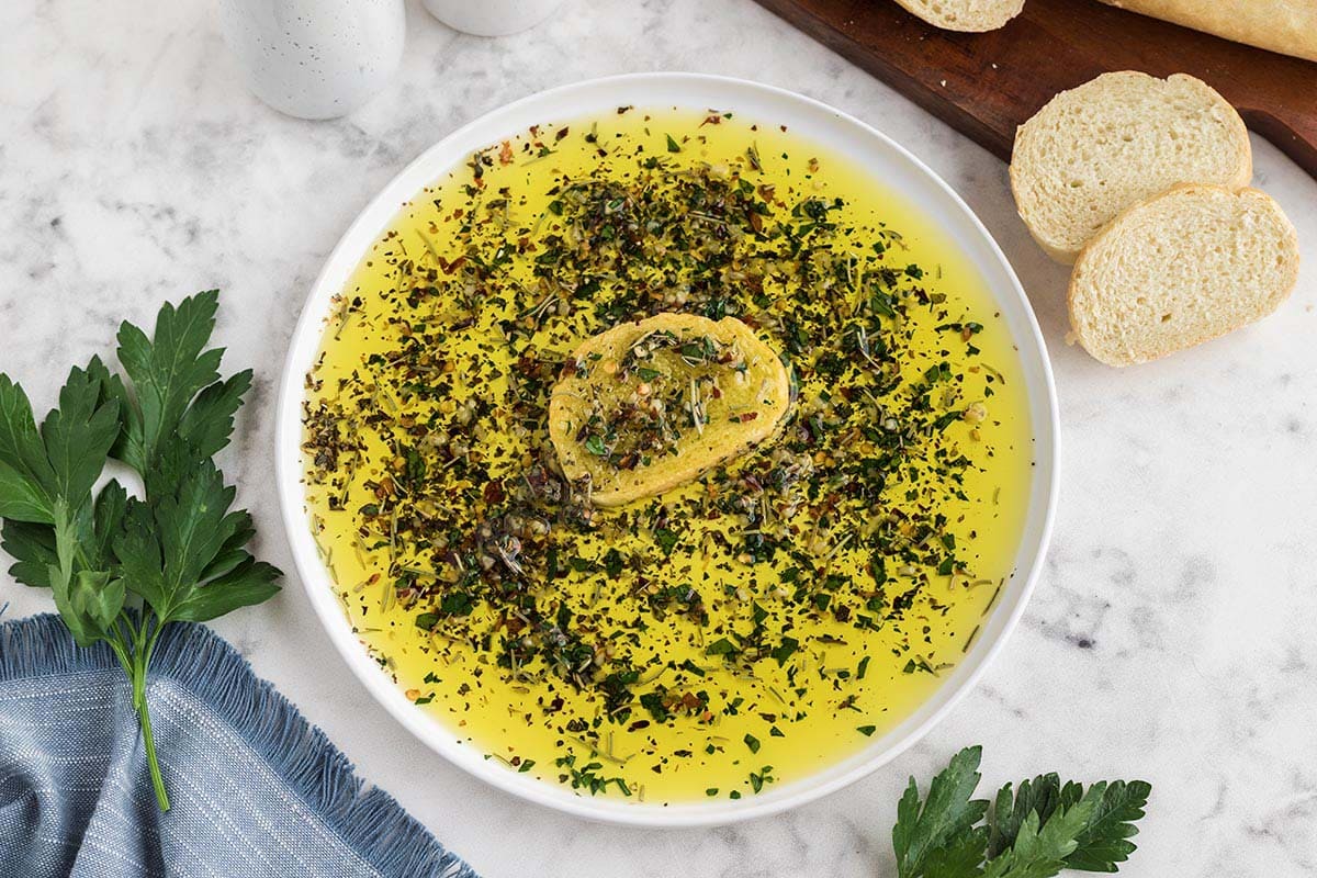White plate filled with olive oil, herbs, and a piece of bread for dipping.