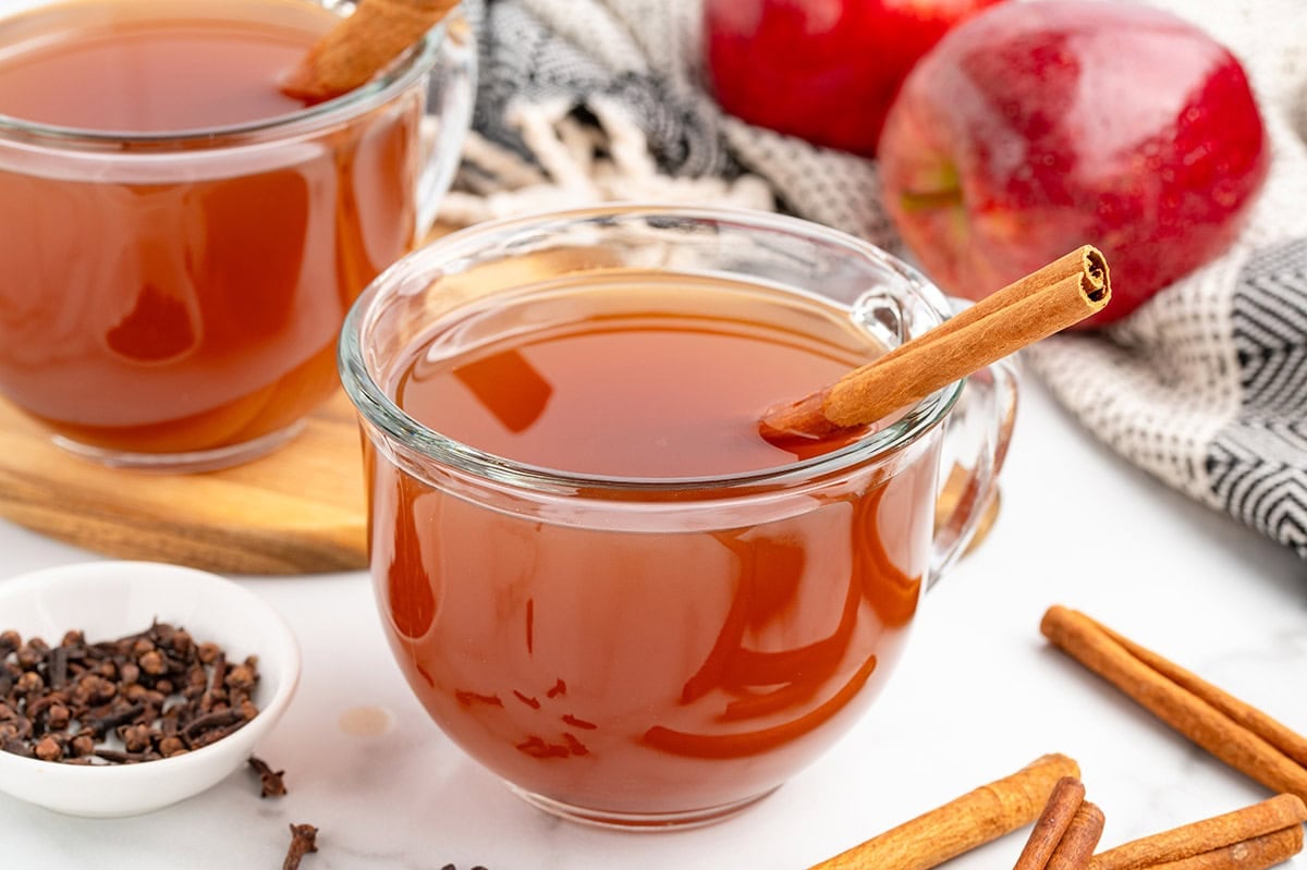 Clear glass cup filled with warm apple cider and a cinnamon stick inside.