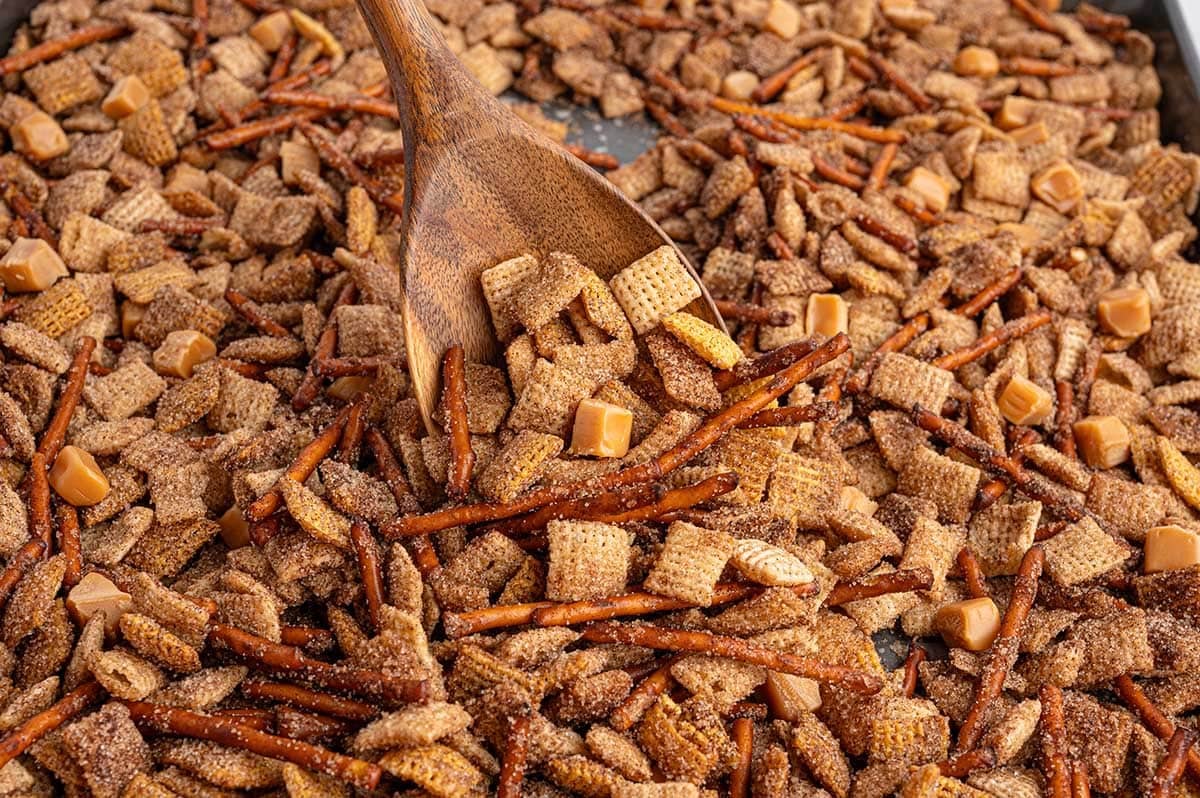 A wooden spoon scooping a batch of cinnamon-coated Chex mix with pretzels and caramel pieces.