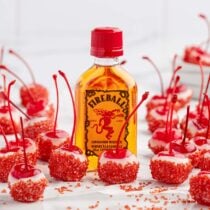 A small bottle of Fireball cinnamon whiskey surrounded by red sugar-coated cherries with stems.