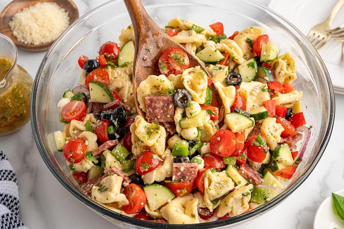 A glass bowl filled with tortellini pasta salad mixed with cherry tomatoes, olives, and cucumbers.
