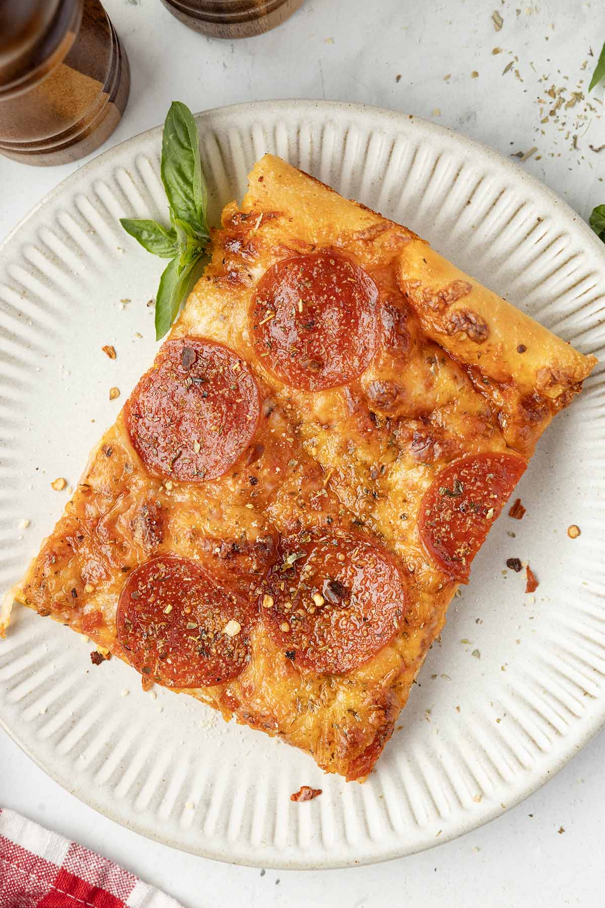 A slice of pepperoni sheet pan pizza on a white plate with a fresh basil leaf.