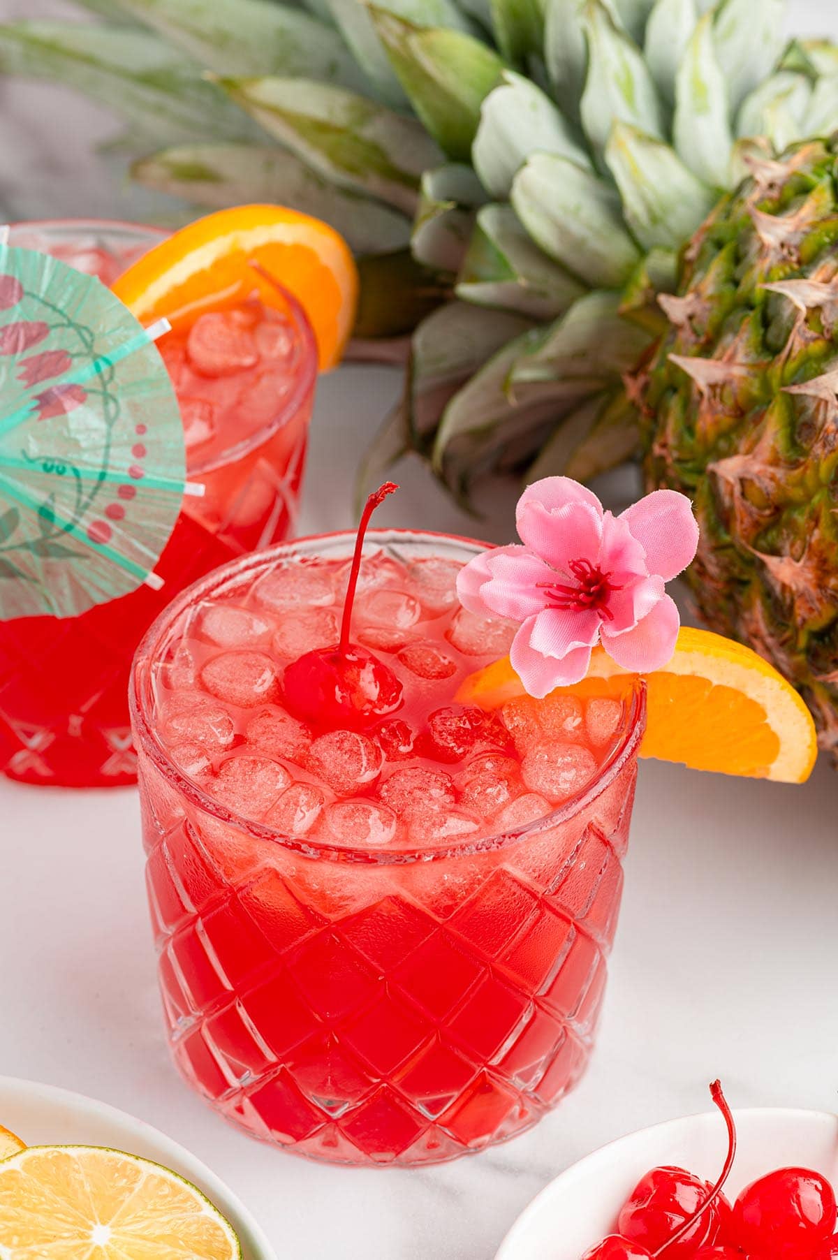 Refreshing red punch in tumblers garnished with cherries orange wedges and pink flowers beside a pineapple.