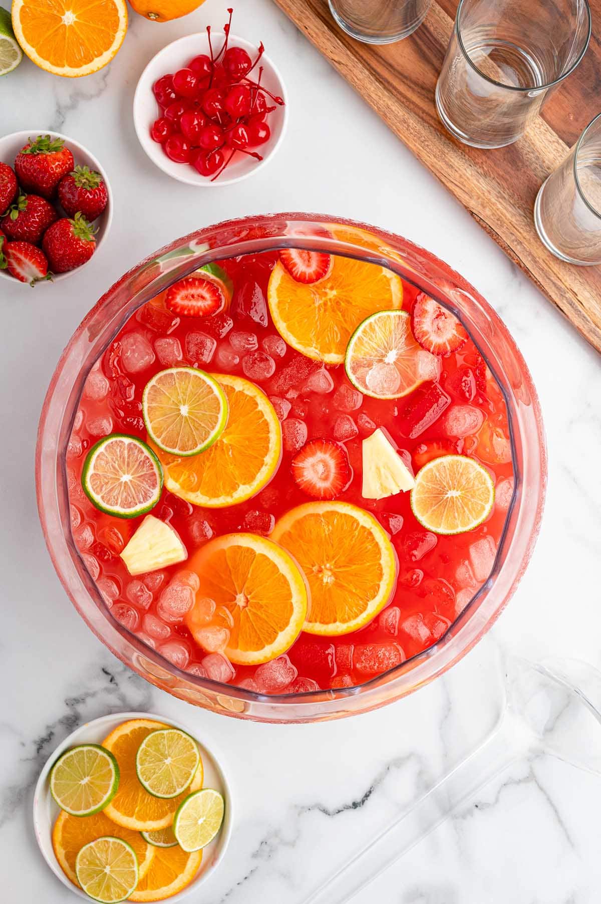 Clear glass bowl of red punch loaded with strawberries, orange segments, lime pieces, pineapple bits, and plenty of ice.