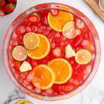 Red party punch in a clear bowl with floating strawberries, orange slices, lime wedges, and pineapple chunks plus ice cubes.
