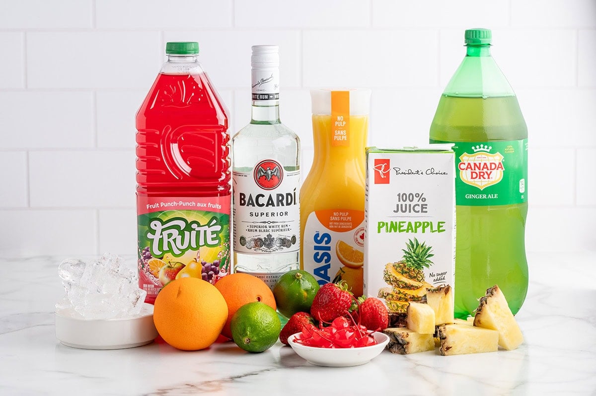 Fresh ingredients for party punch: Bacardi rum, cranberry juice, orange juice, pineapple juice, and tropical fruits like oranges, limes, strawberries, cherries, and pineapple chunks on a white table with ice.
