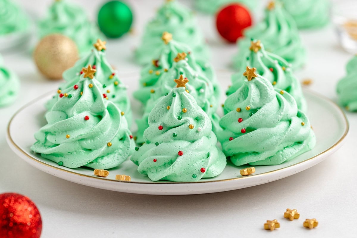 Green christmas tree meringue cookies sit on a white plate with gold stars and colorful ornaments nearby.