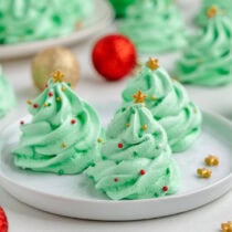 Green meringue cookies shaped like Christmas trees sit on a white plate with red and gold ornaments nearby.