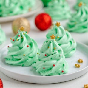 Green meringue cookies shaped like Christmas trees sit on a white plate with red and gold ornaments nearby.