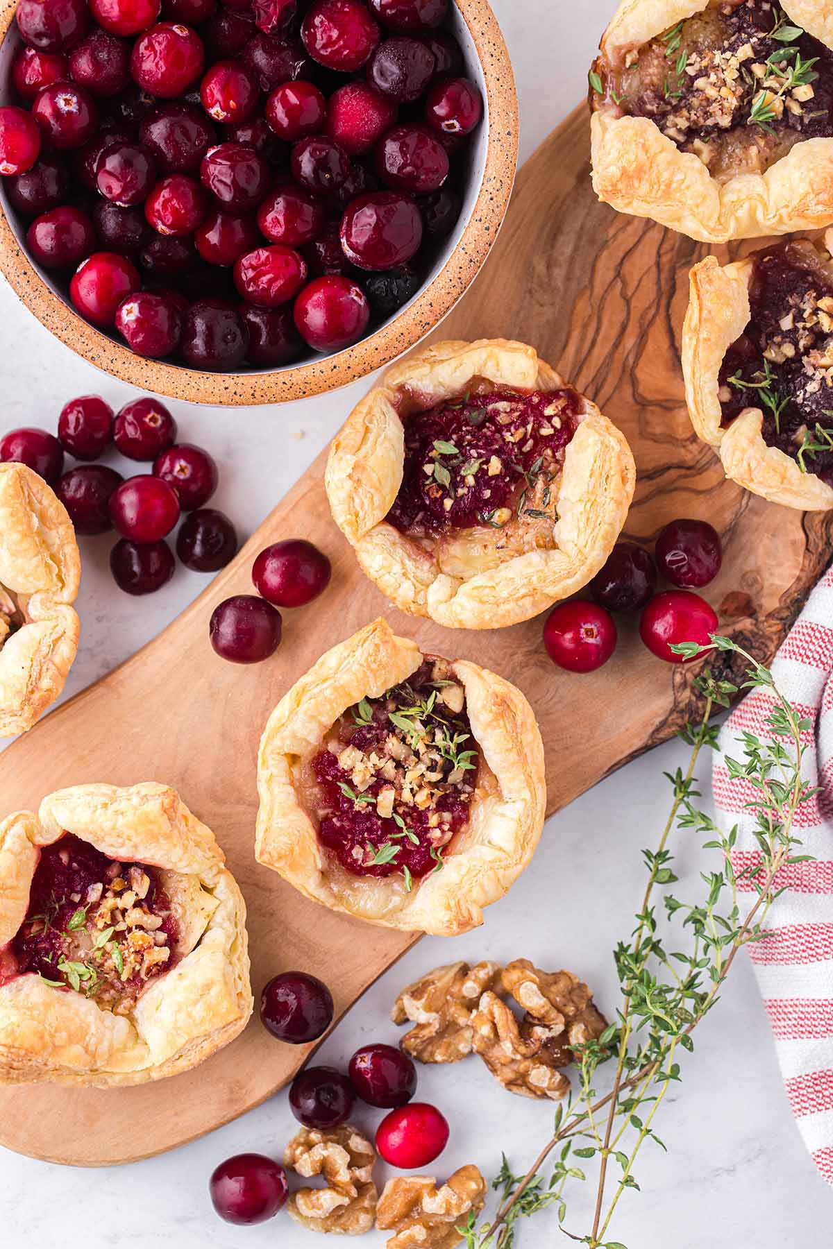Fresh cranberries fill a bowl next to golden brie bites topped with nuts on a wooden board.