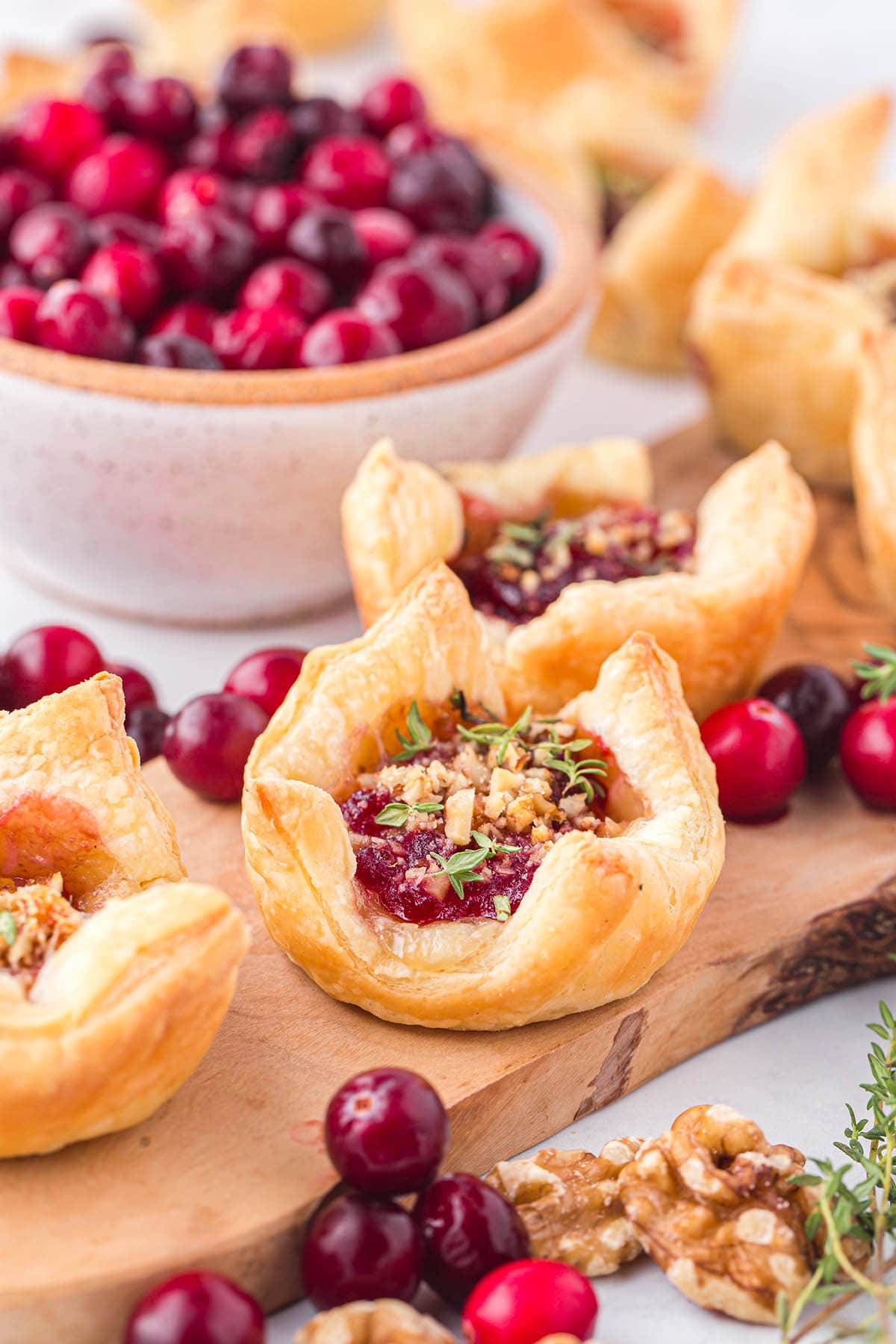 Fresh cranberries in a bowl next to baked brie bites on a wooden board, with scattered nuts and herbs.