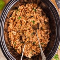 Crockpot filled with seasoned shredded chicken and two serving spoons.
