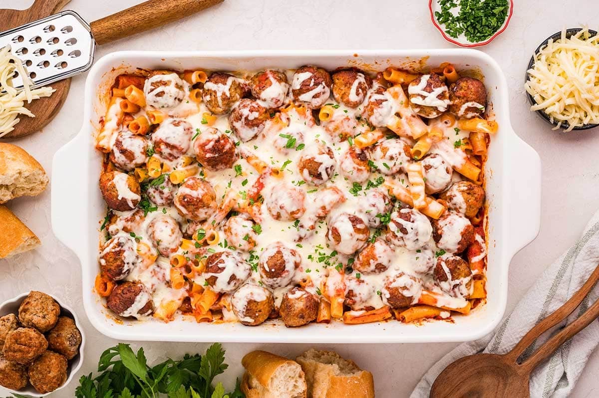 Meatball casserole in a dish, surrounded by baguette slices and a wooden spoon.