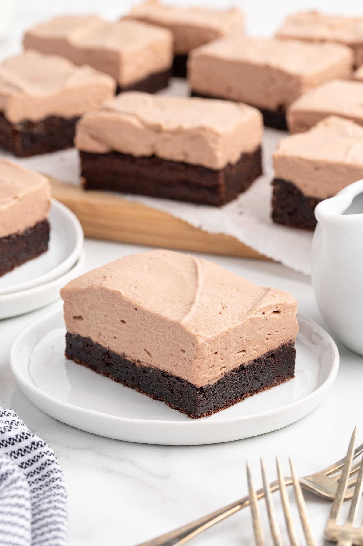 Square shaped chocolate mousse brownies with smooth tan mousse topping sits on a white plate.