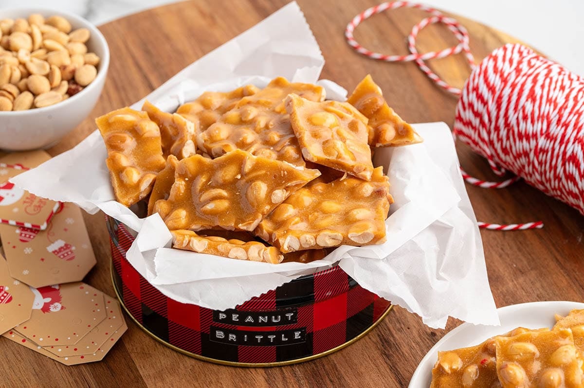 A red and white checkered tin holds fresh peanut brittle pieces wrapped in white paper.