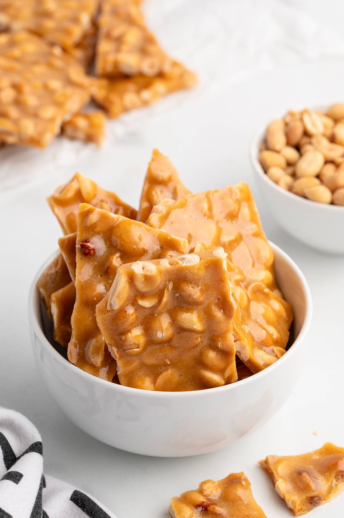 A white bowl holds golden peanut brittle with whole peanuts mixed in