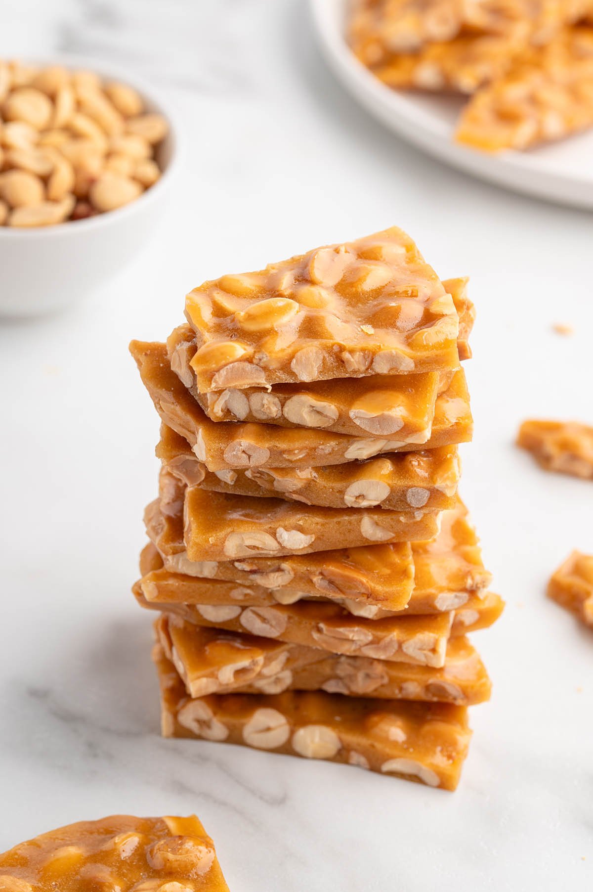 A tall stack of golden peanut brittle sits on a white surface next to a bowl of peanuts.