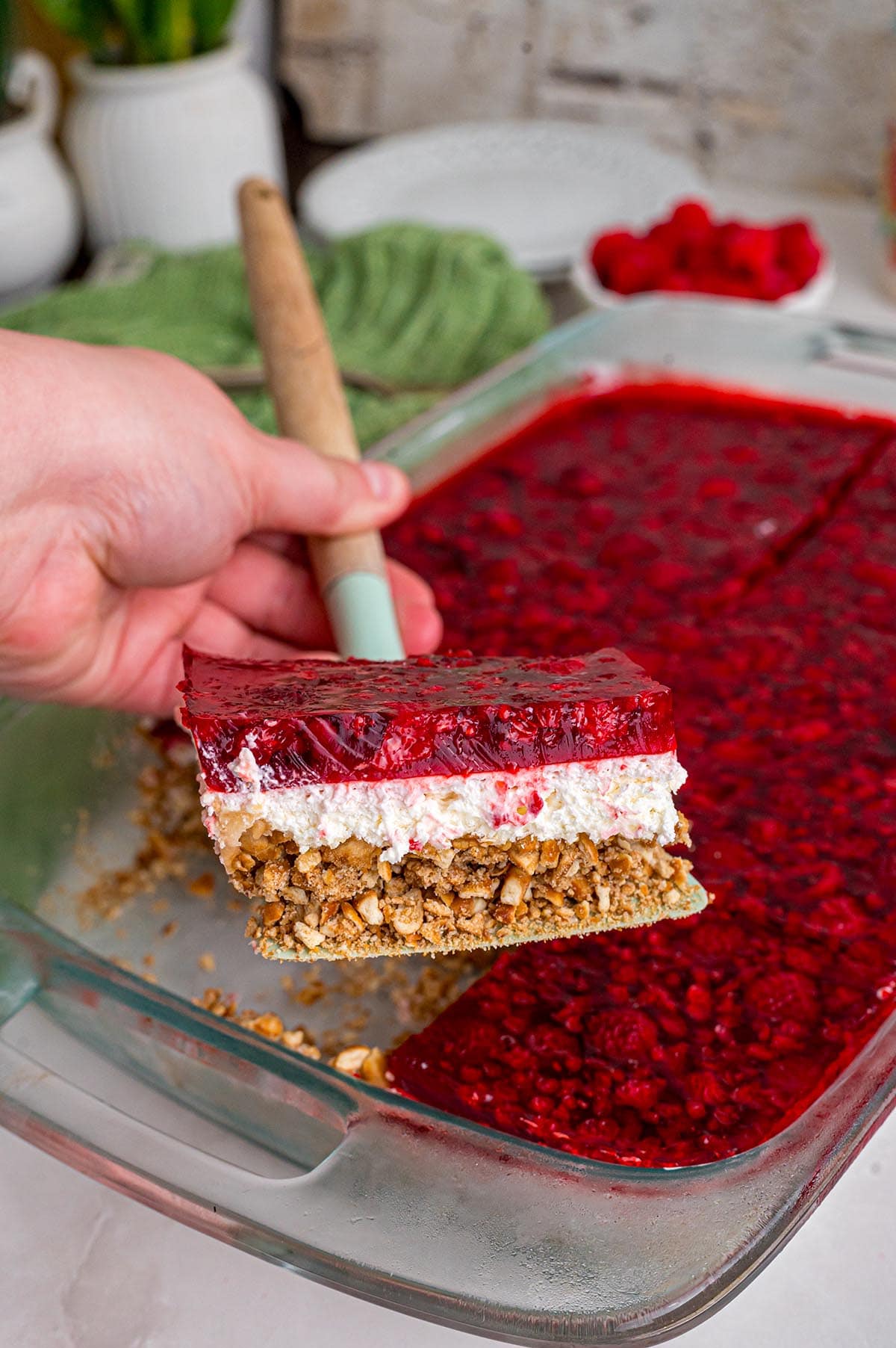 Hand holding a serving spoon lifting a piece of raspberry pretzel salad from a glass baking dish.