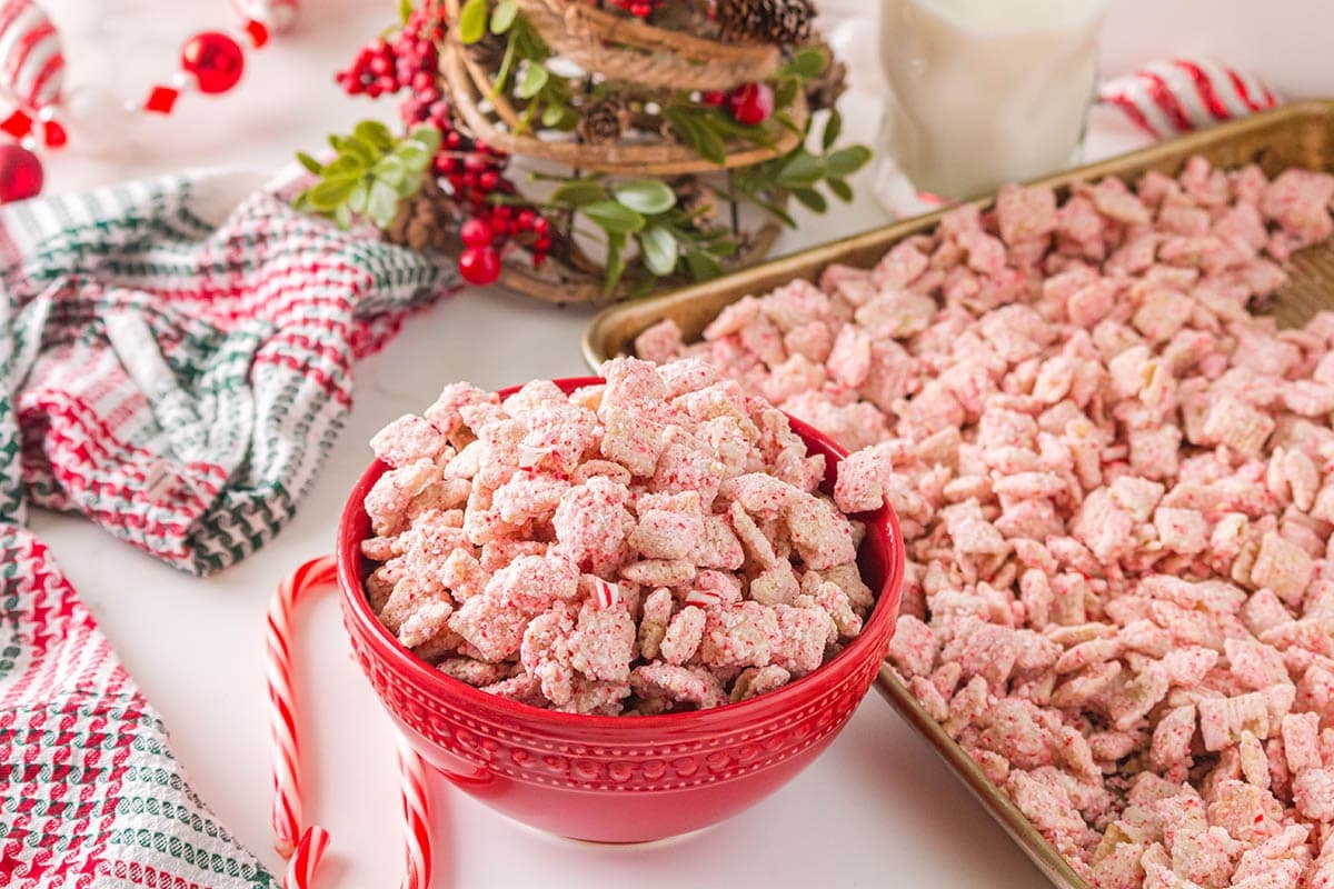 Pink chunks of candy cane muddy buddies fill a red bowl, surrounded by holiday garland and a candy cane.