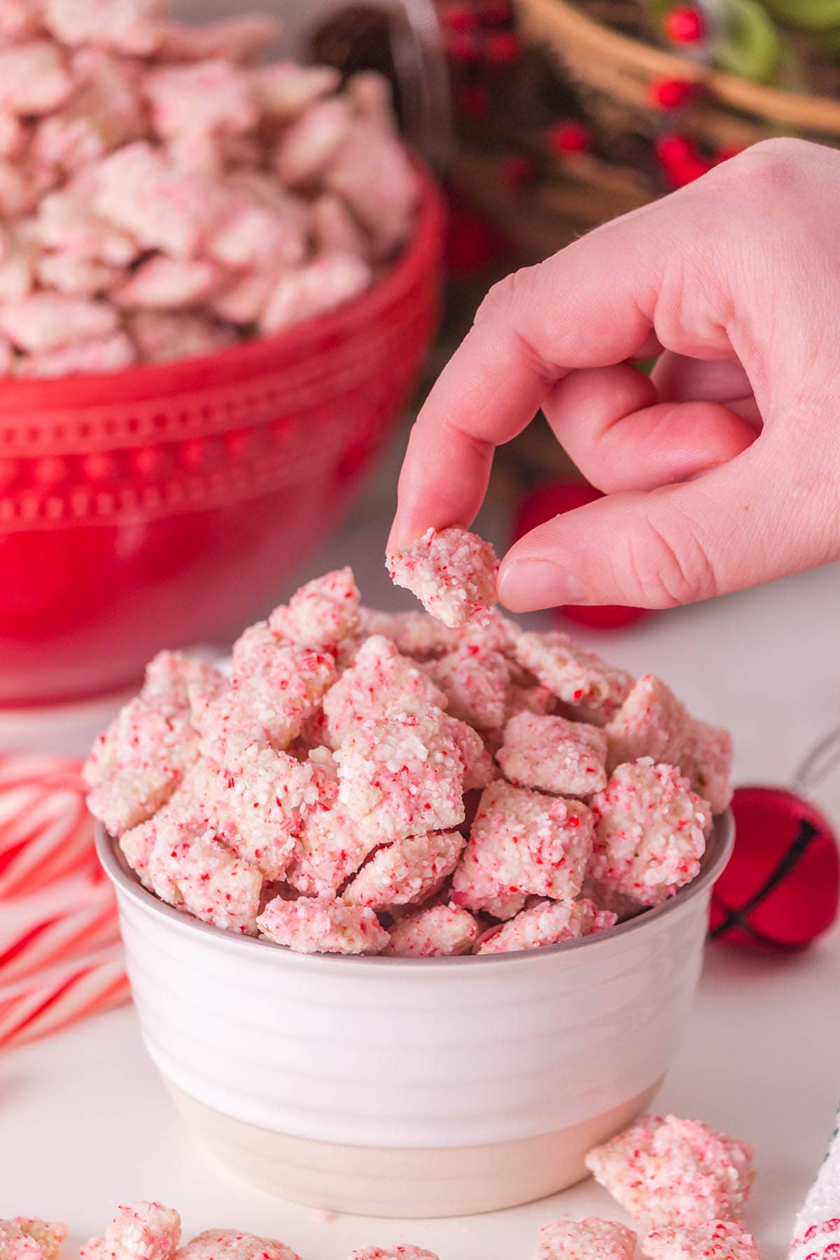 A white bowl holds pink candy cane muddy buddies Chex mix, with a hand grabbing a crunchy piece.