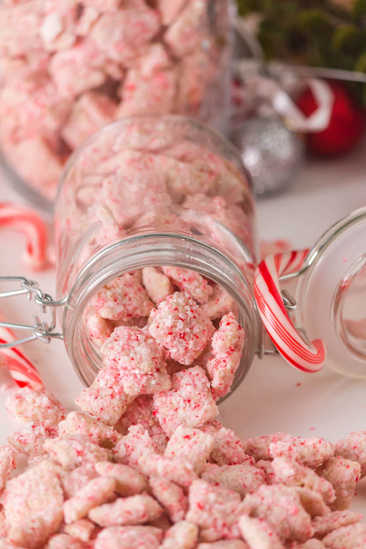 A clear glass jar holds pink candy cane muddy buddies Chex mix, spilling out onto a white table.