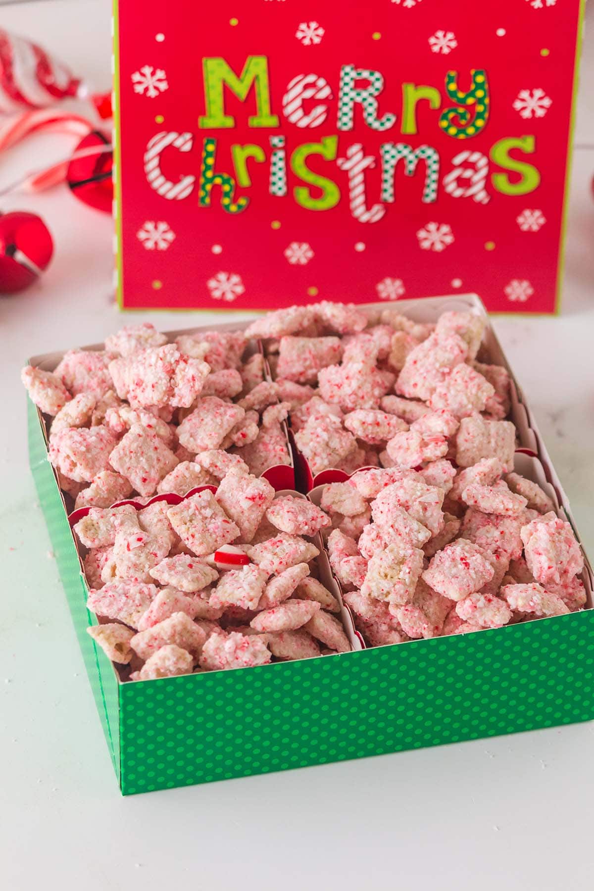Pink candy cane muddy buddies Chex mix fills a green box, with a red Merry Christmas gift box nearby.