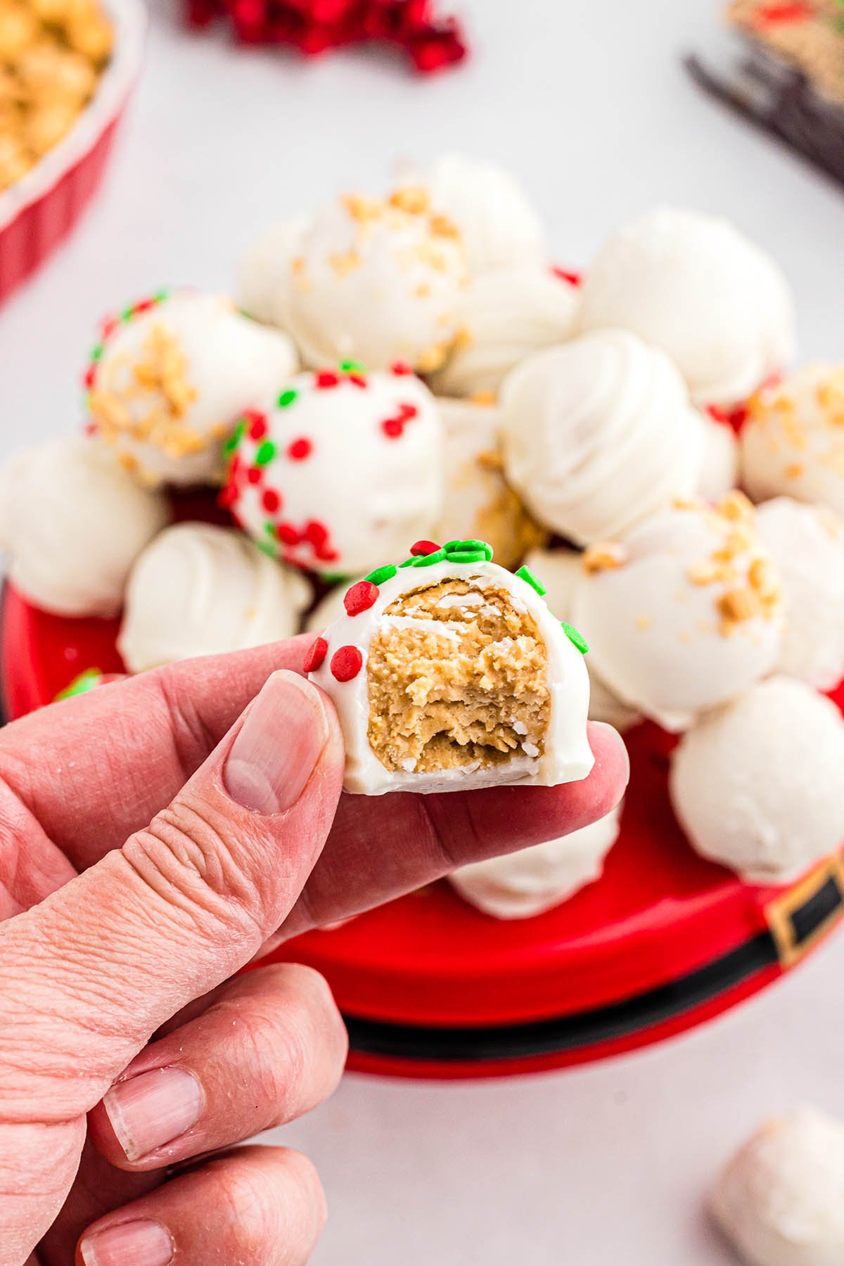 A hand holds a bitten white chocolate truffle with peanut butter filling, surrounded by holiday truffles in red and green sprinkles.