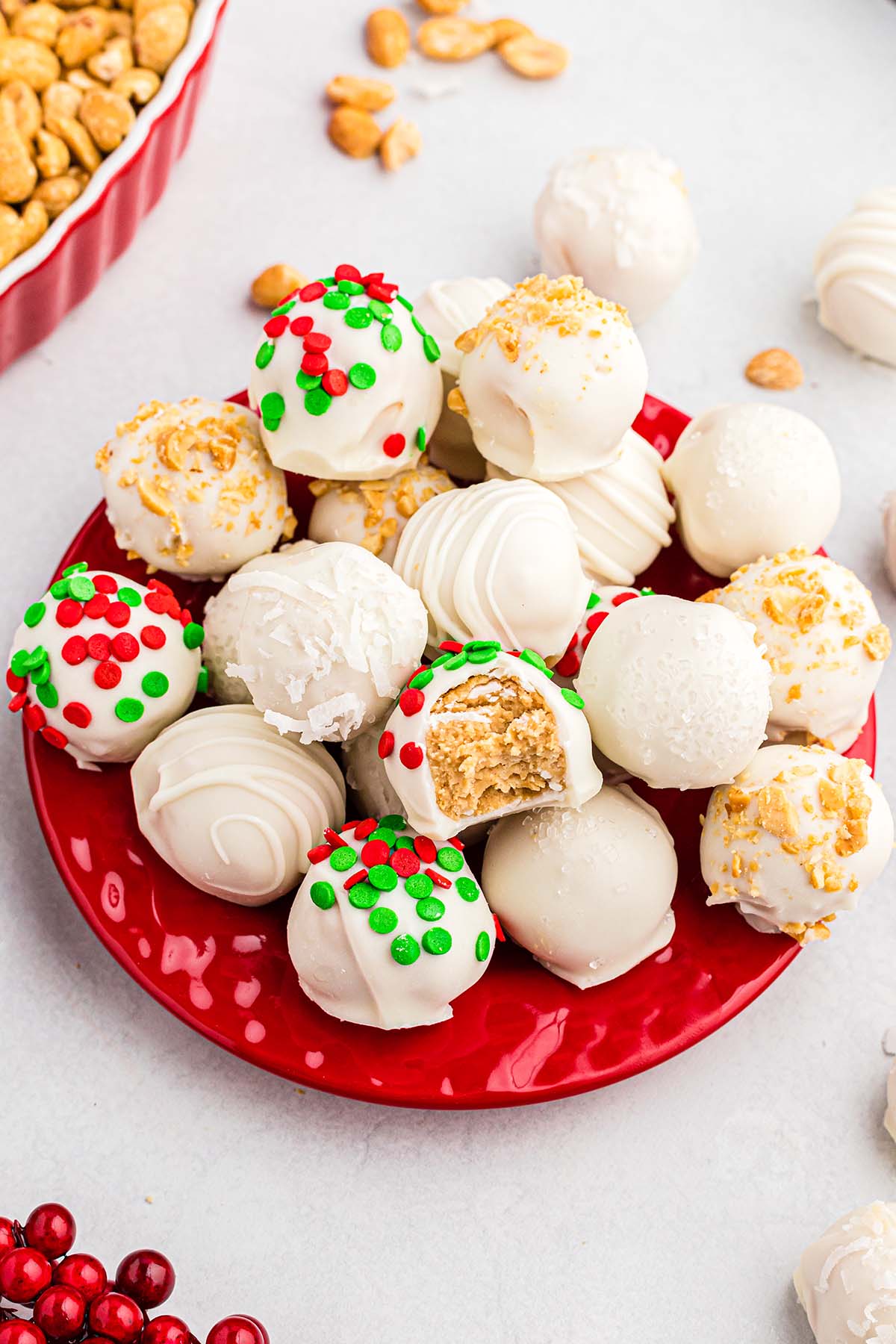 Festive Christmas truffles sit on a red plate, featuring white chocolate balls with coconut and colorful sprinkles, plus fresh cranberries nearby.