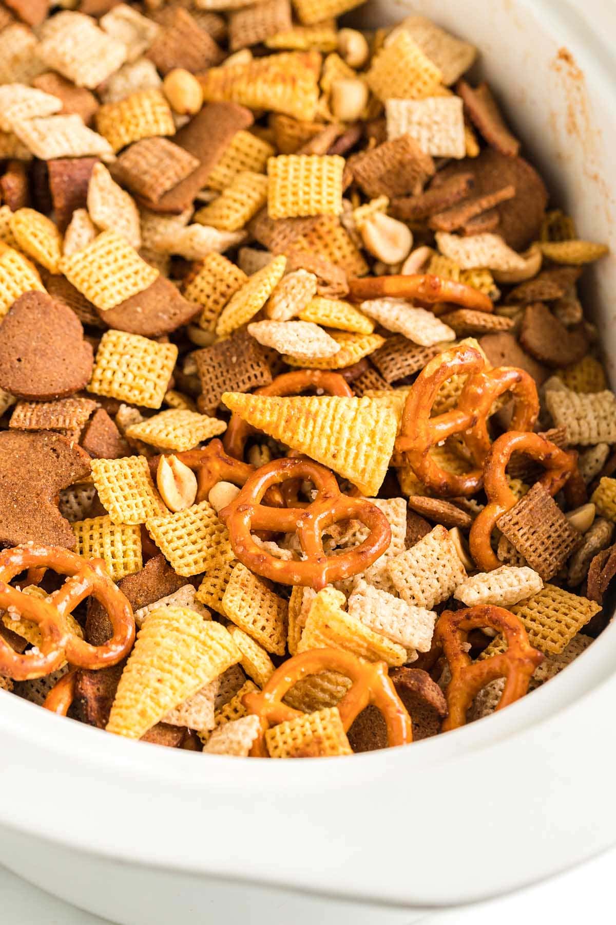A white crockpot brims with homemade Chex Mix, featuring square cereals, pretzels, and peanuts.