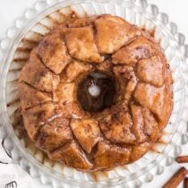 Golden monkey bread sits on a clear plate with shiny glaze and cinnamon sticks nearby.