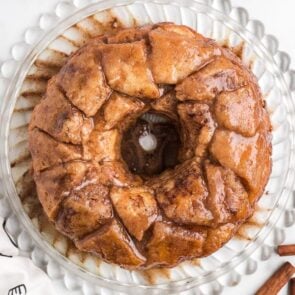 Golden monkey bread sits on a clear plate with shiny glaze and cinnamon sticks nearby.