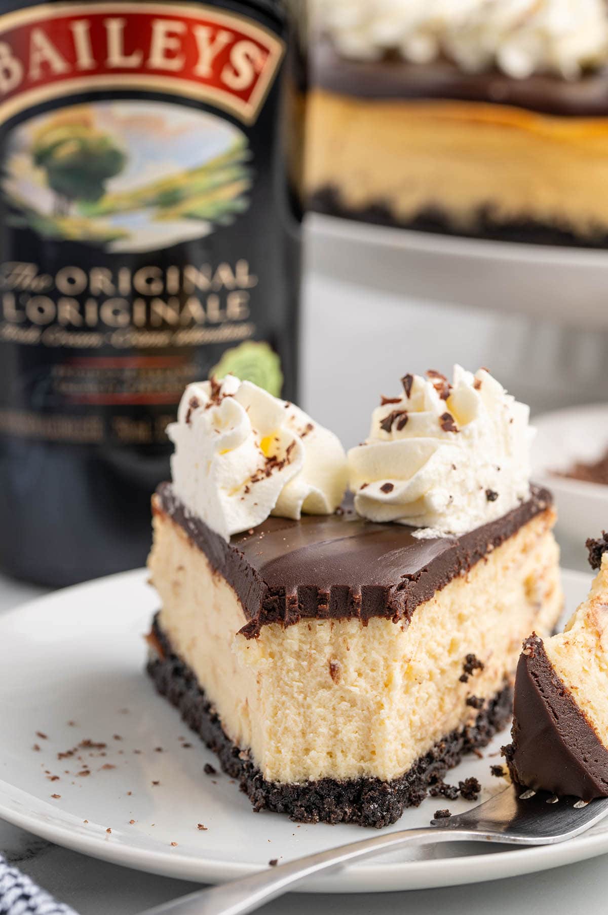 Slice of creamy Bailey's cheesecake topped with chocolate ganache and whipped cream on a white plate.