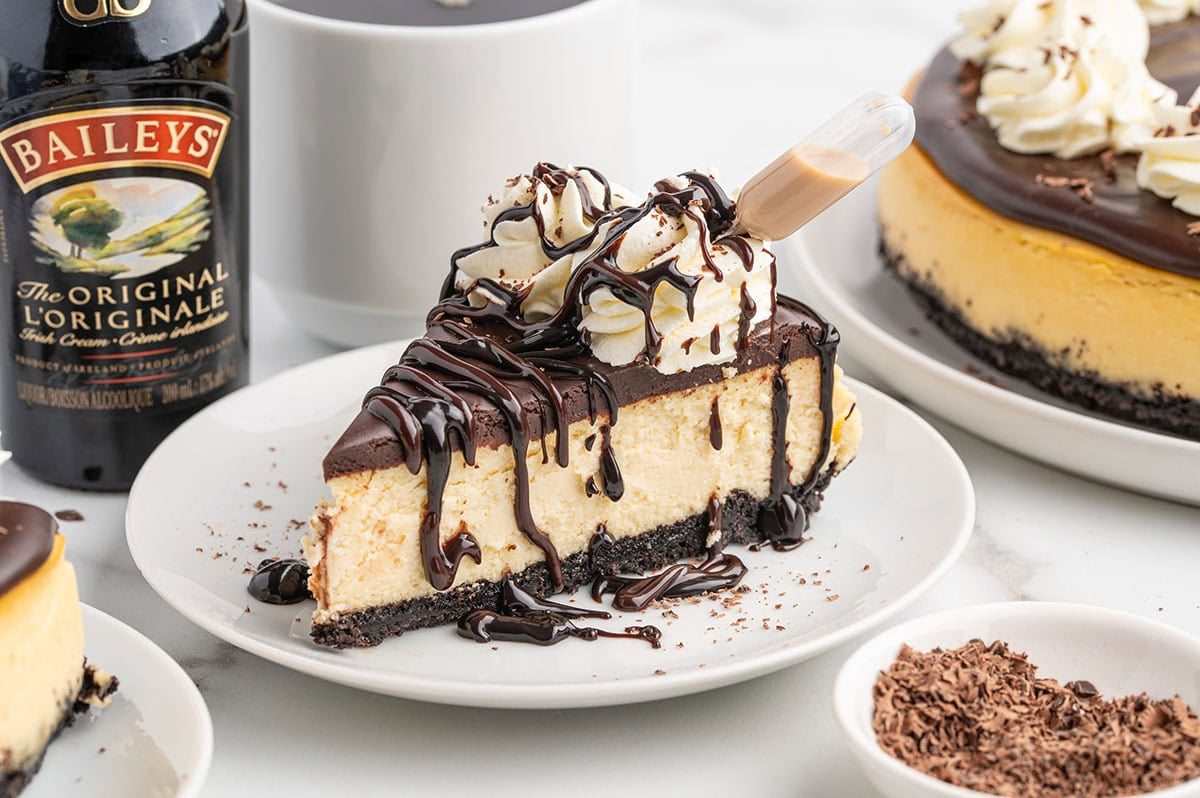 A slice of Baileys cheesecake drizzled with chocolate sits on a white plate next to a bottle of Baileys Irish Cream.