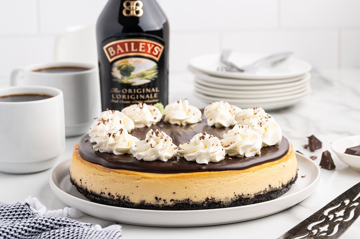 A slice of Bailey's cheesecake topped with chocolate ganache and whipped cream sits on a white plate next to a bottle of Baileys Irish Cream.