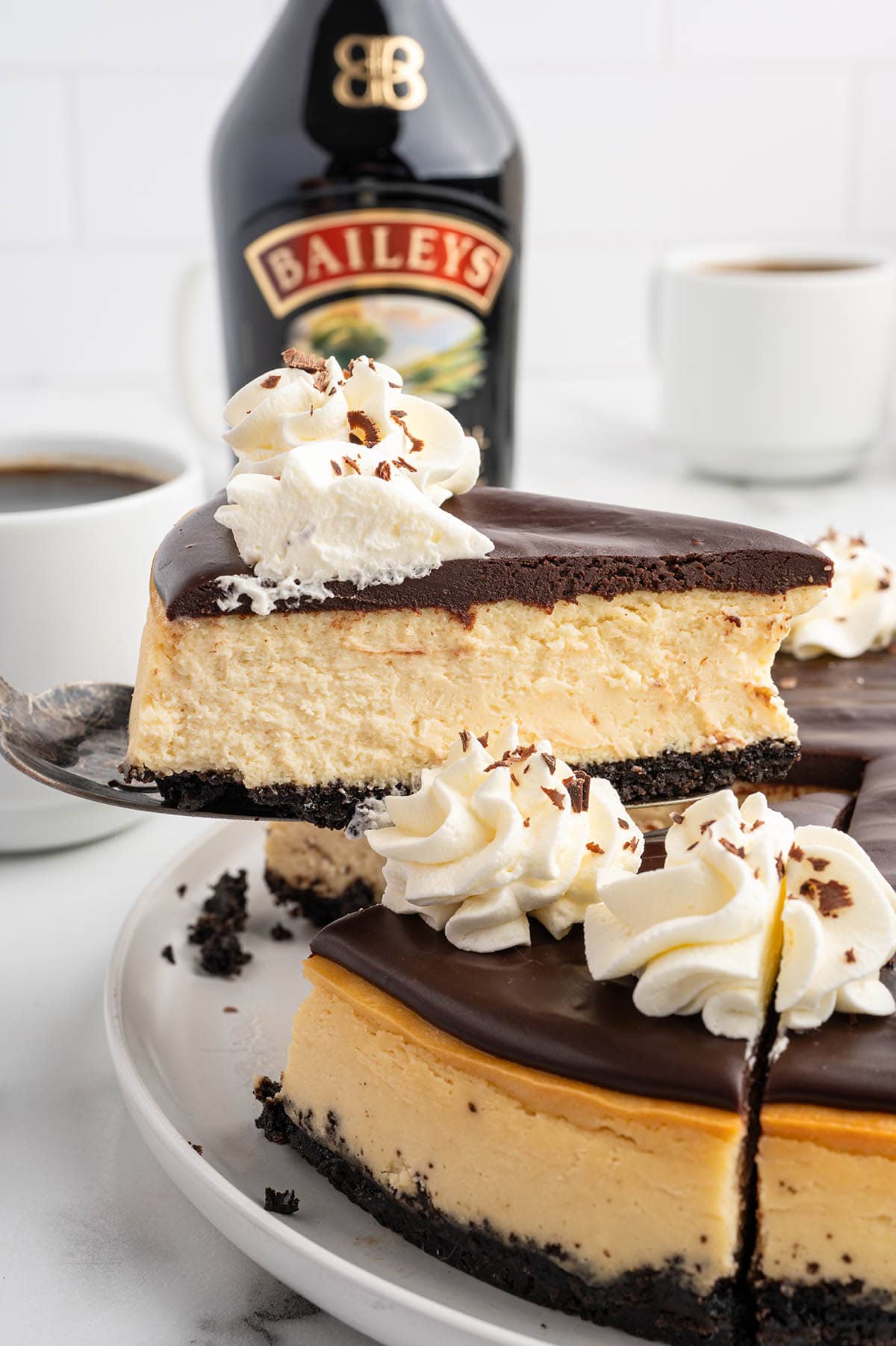 A creamy slice of Baileys cheesecake with chocolate topping and whipped cream sits on a plate next to a bottle of Baileys and a cup of coffee.
