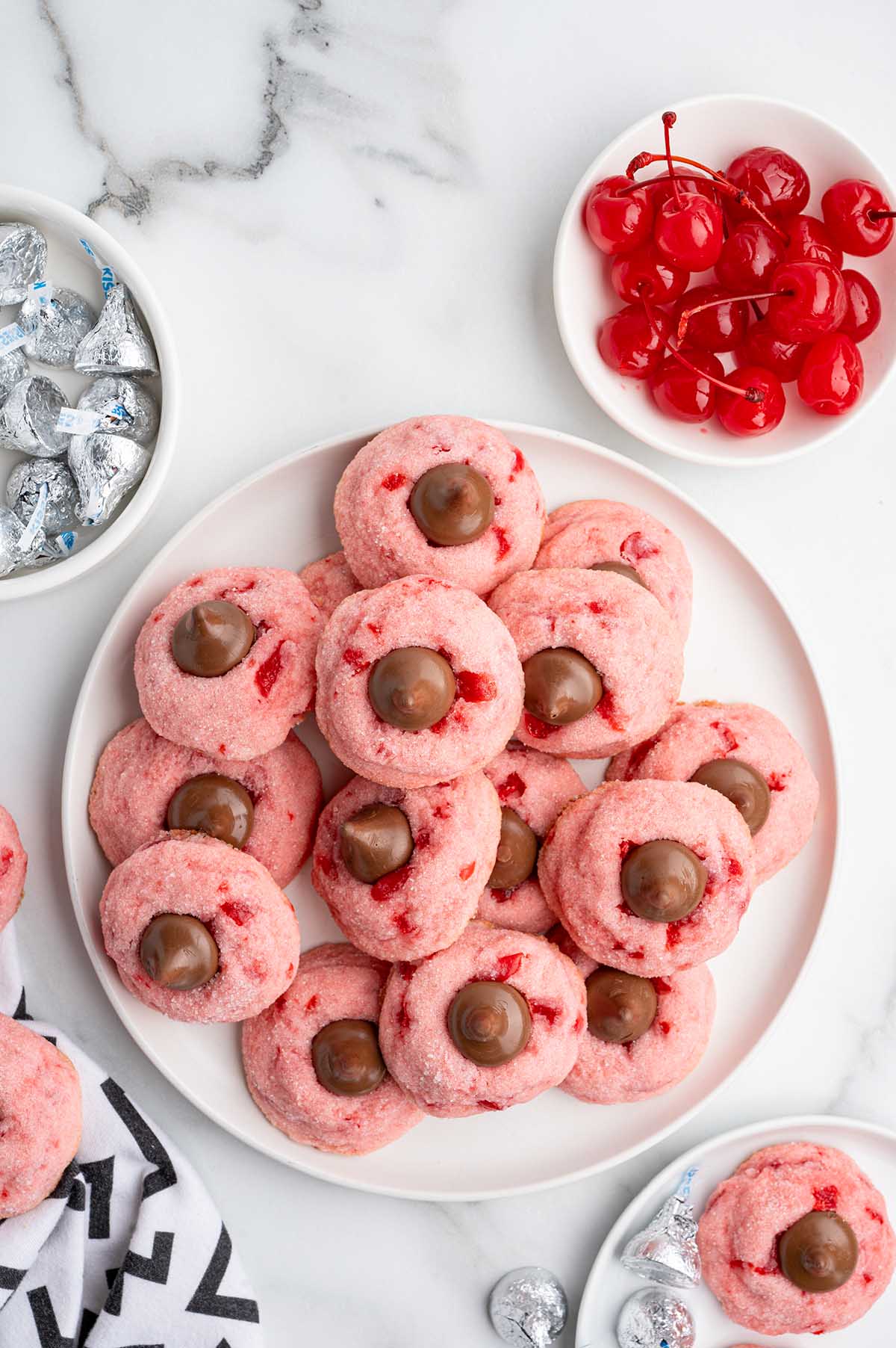 Pink cherry kiss cookies topped with chocolate bits sit on a white plate next to bowls of cherries and Hershey's kisses.