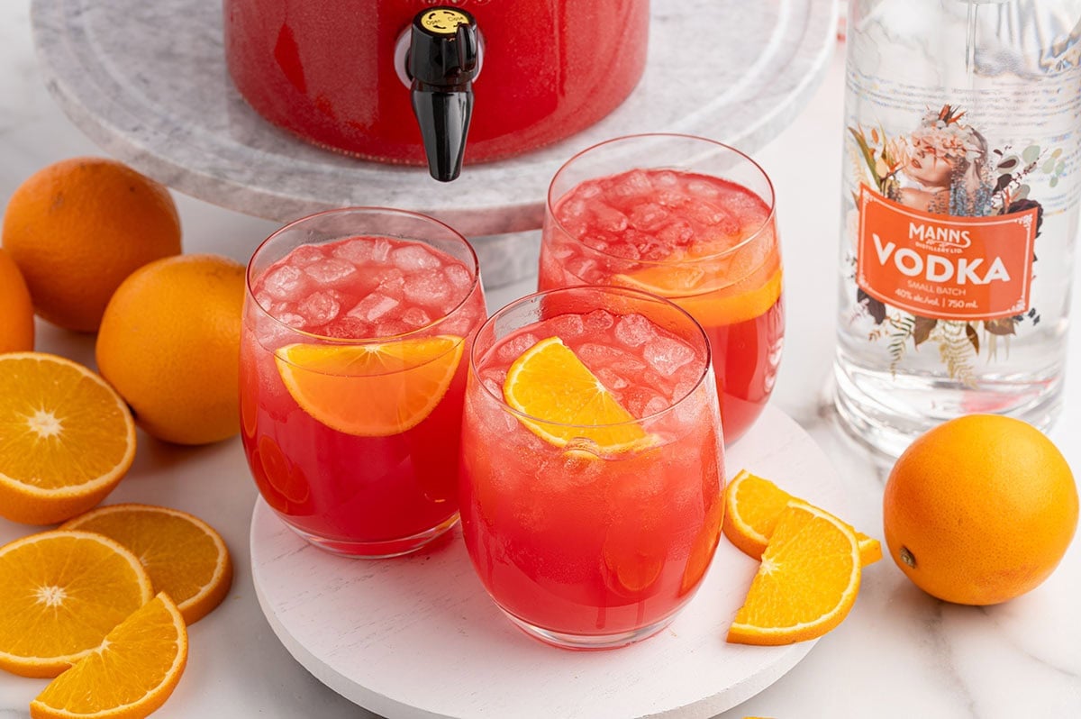 Three glasses of red Christmas Morning Punch with ice and orange slices sit next to a vodka bottle and fresh oranges.