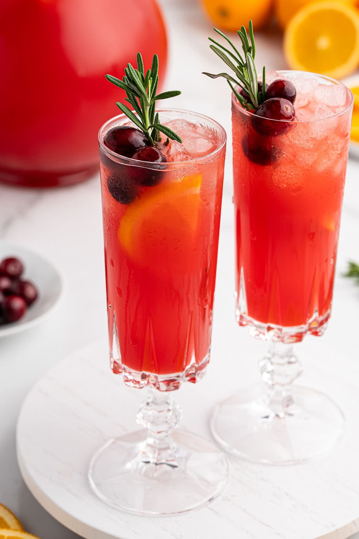 Two tall glasses of red Christmas Morning Punch topped with cranberries, orange slices, and rosemary sprigs sit on a white surface.