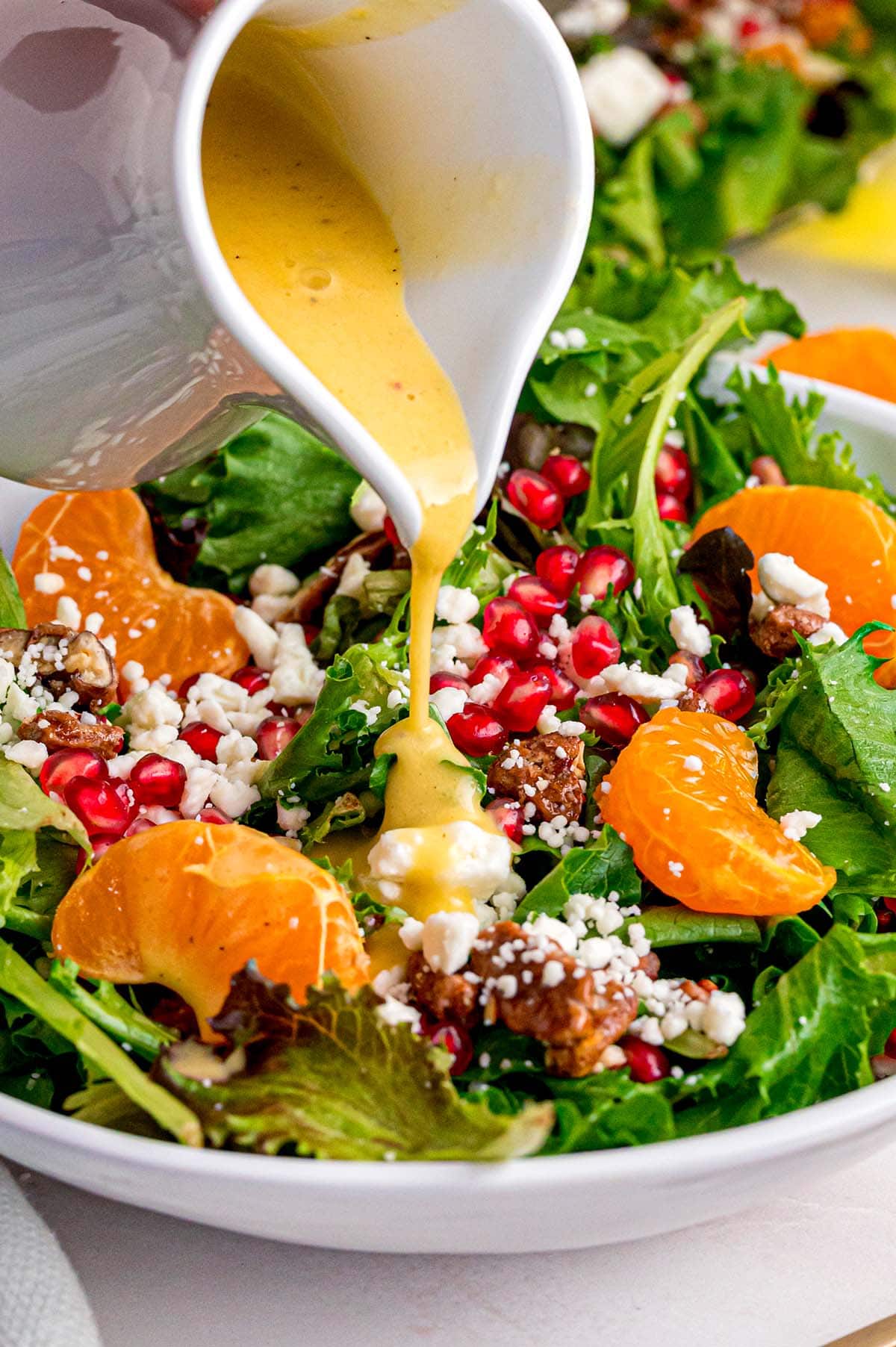 A hand pours creamy yellow dressing over a fresh salad bowl filled with mixed greens, mandarin orange slices, pomegranate seeds, feta cheese, and walnuts.