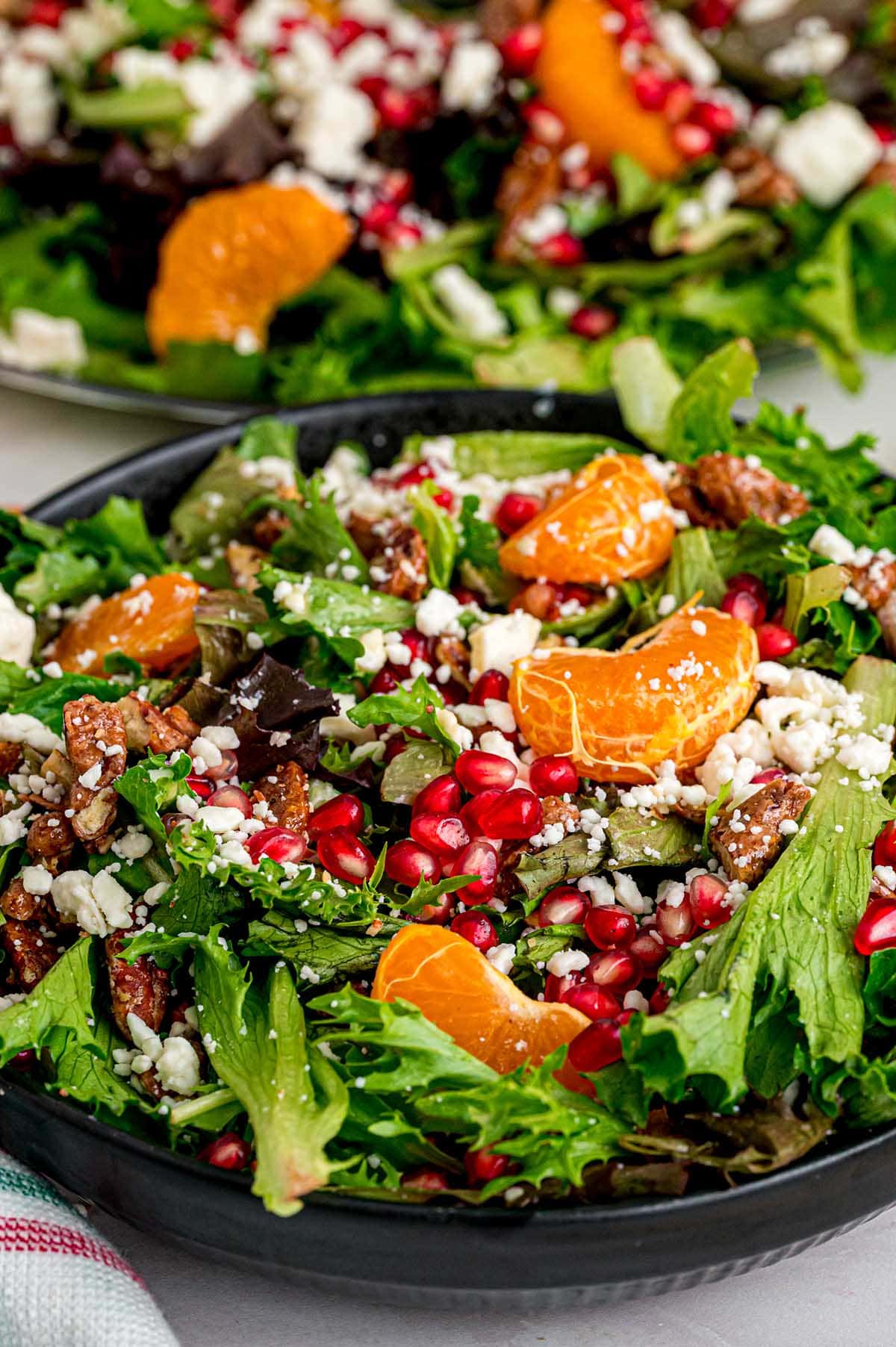 Fresh Christmas salad in a black bowl with greens, orange segments, pomegranate seeds, feta cheese, and walnuts.