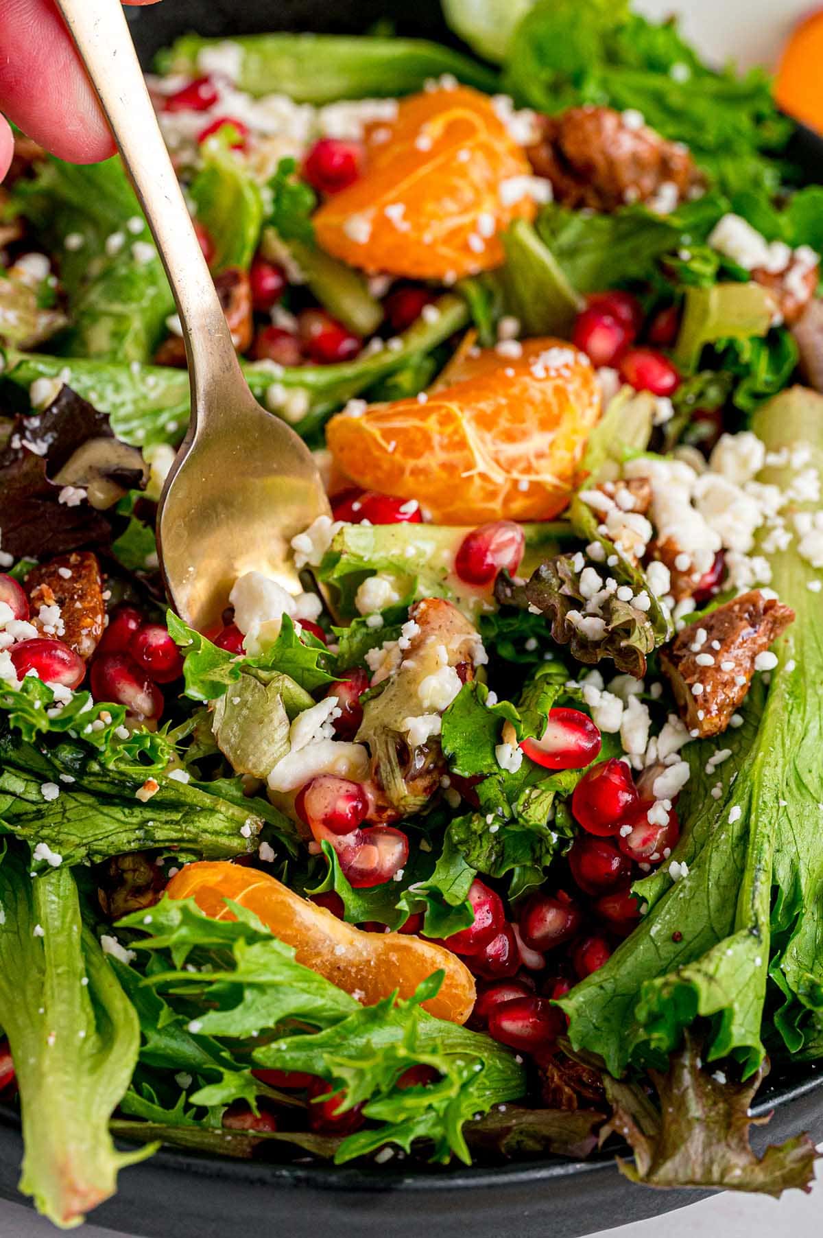 A hand lifts a spoonful of fresh Christmas salad from a black bowl, showing green lettuce, red pomegranate seeds, orange slices, white feta, and nuts.