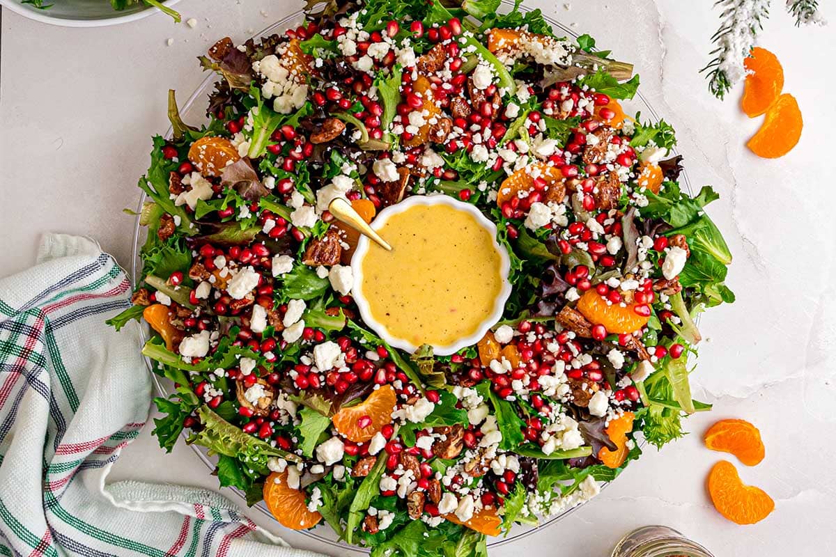 A vibrant Christmas salad shaped like a wreath, topped with feta, pomegranate seeds, mandarin slices, and pecans, with dressing in the center.