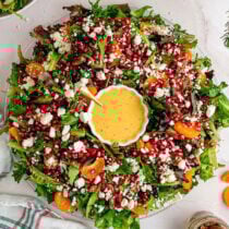 Festive Christmas salad wreath with fresh greens, red pomegranate seeds, white feta crumbles, orange mandarin slices, and pecans around a bowl of creamy dressing.